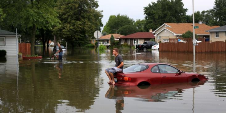 A man sitting on top of car in the middle of flooded area.